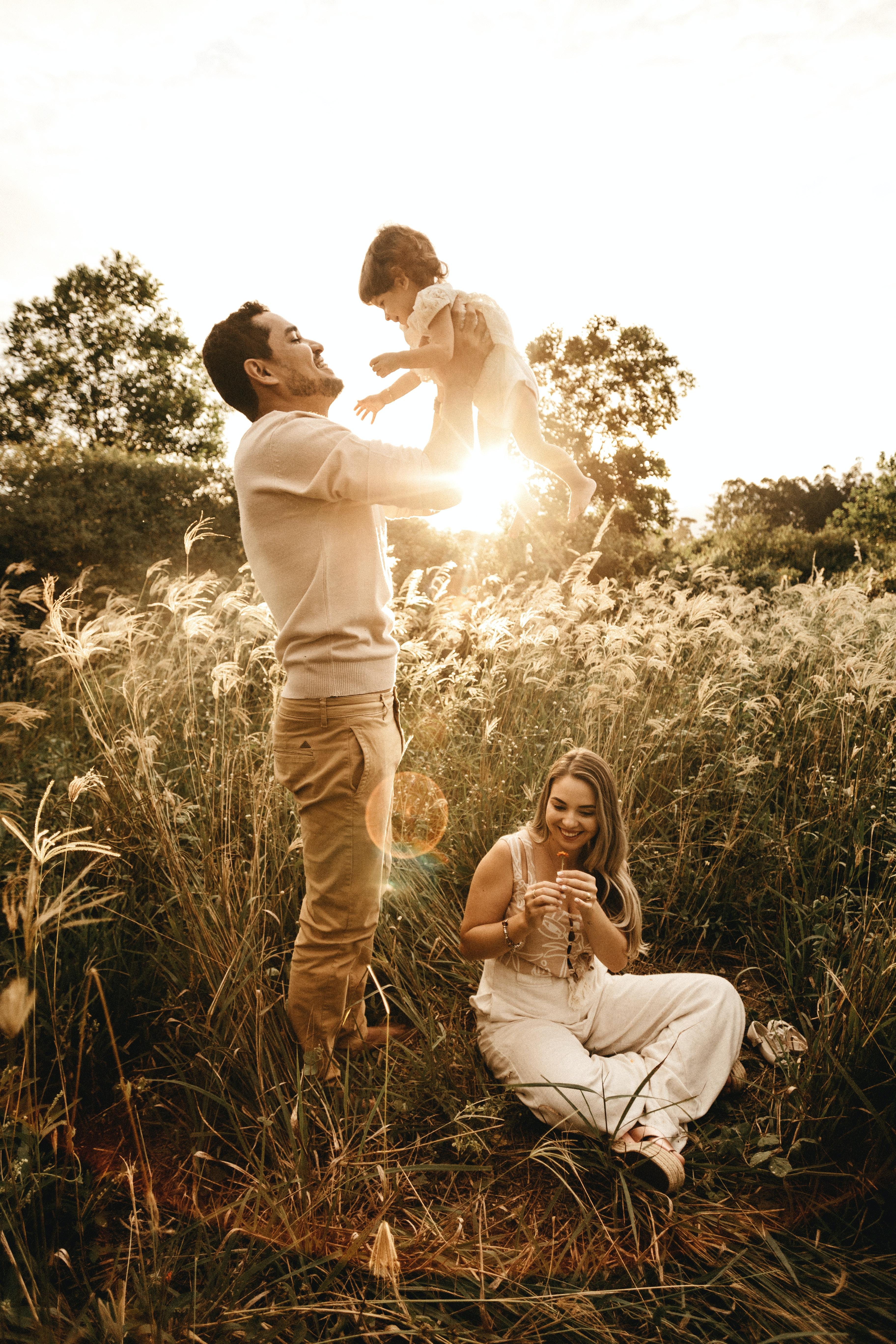 family in grass field. Father holding baby up. Mother sitting on ground